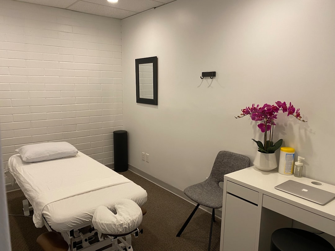 A minimalist massage room with a massage table, a chair, and decorative orchids on a white desk